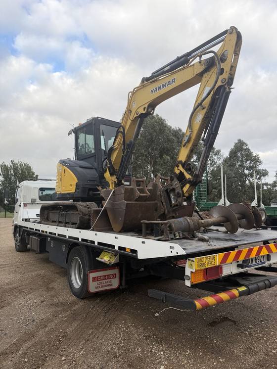 Hilly's truck carrying a yellow excavator with buckets and an auger attachment